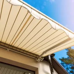 Toldo beige desplegado sobre terraza, cumpliendo la normativa. Cielo azul y árbol al fondo.