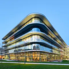Edificio moderno con **cristales curvos** que reflejan el cielo azul. Fachada ondulada y luminosa, con áreas verdes y árboles alrededor.