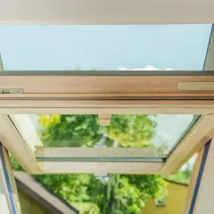 Ventana de tejado abierta, mostrando el marco de madera y el cristal con cámara de aire. Se ve el cielo azul y árboles verdes al exterior.