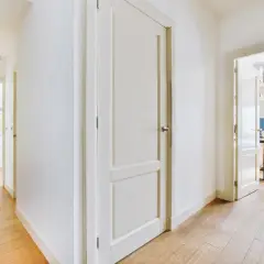 Pasillo con puertas de interior blancas y suelo de madera. Se vislumbran una cocina y un despacho.