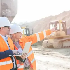 Dos ingenieros con cascos y chalecos naranjas revisan un plano en una obra. Uno señala al fondo, donde se ve una excavadora.