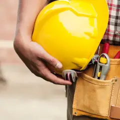Un trabajador sostiene un casco amarillo. Su cinturón de herramientas tiene destornilladores y una llave inglesa. Esto es lo que es el ITE: seguridad y preparación.