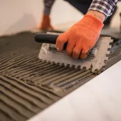 Man con guantes naranjas esparce mortero con llana dentada, preparando el suelo para colocar baldosas.