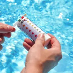 Manos sostienen una tira reactiva para medir el pH de la piscina. El agua azul y cristalina de la piscina se ve al fondo.