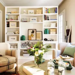 Acogedor salón con estanterías blancas llenas de libros y objetos decorativos, dos sillones de rayas y una mesa de centro con flores. Un rincón que evoca la calidez de las casas pequeñas.