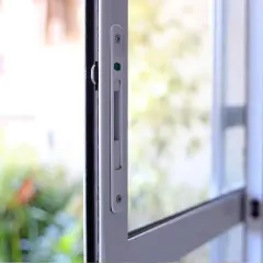 Detalle de un mecanismo de cerraduras seguras en una ventana corrediza, con un fondo desenfocado de vegetación y luz.