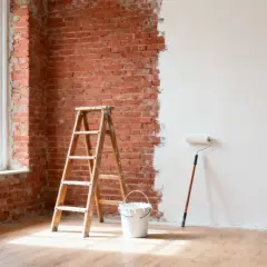 Una habitación con paredes de ladrillo expuesto y una pared parcialmente pintada de blanco. Una escalera de madera, un cubo de pintura y un rodillo esperan para continuar la renovación.