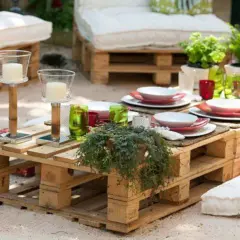 Mesa de jardín con ideas con palets, decorada con vajilla, velas y plantas. Cojines a los lados invitan a relajarse.