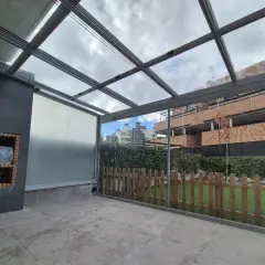 Patio con techo de cristal y estructura metálica, ideal para techar patio económico. Incluye barbacoa y valla de madera.