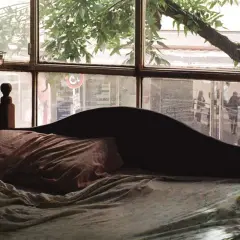 Dormitorio con ventana en el cabecero. La cama está hecha, con sábanas arrugadas y una almohada. A la izquierda, un mueble de madera con cajones.
