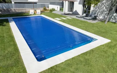 Piscina cubierta con persiana azul, lista para climatizar y disfrutar. Césped verde y tumbonas invitan al relax.