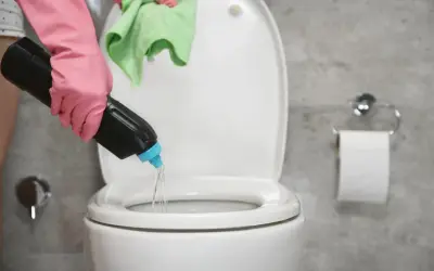 Mano con guante rosa vierte agua fuerte para que sirve en el inodoro. Paño verde en mano.