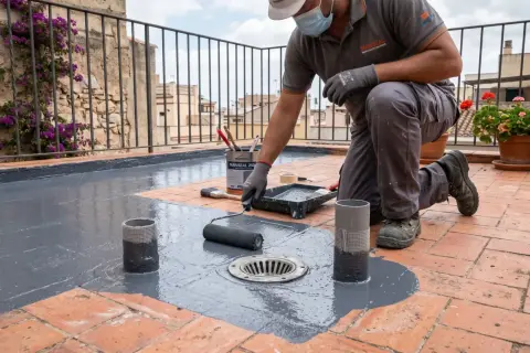Obrero aplica pintura gris para impermeabilizar terraza. Rodillo y cubeta de producto a su lado.