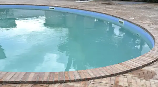 Agua de piscina blanquecina, con reflejos del cielo y rodeada por un borde de ladrillos.