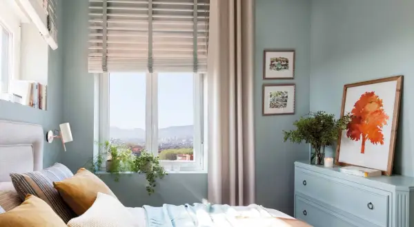 Acogedor plano dormitorio 9 metros cuadrados con cama, cómoda azul, cuadros y vistas a la ciudad.