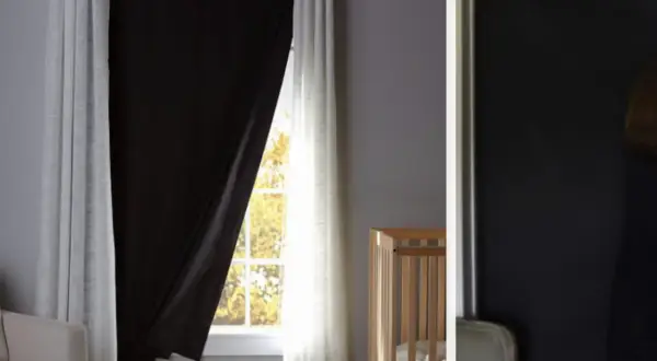Habitación infantil con cuna y una ventana sin persiana, cubierta por cortinas blancas y un panel negro para oscurecer.