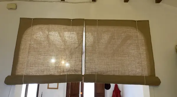 Dos estores enrollables caseros de tela de yute cubren una ventana. La luz del sol se filtra a través de ellos.