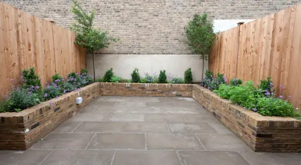 Un patio moderno con jardineras de ladrillo para decorar poyete ventana exterior. Plantas verdes y flores moradas crean un oasis urbano.