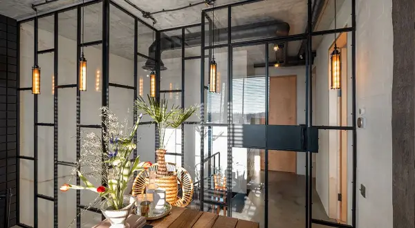 Interior moderno con paredes de cristal, lámparas colgantes y un arreglo floral sobre una mesa de madera.
