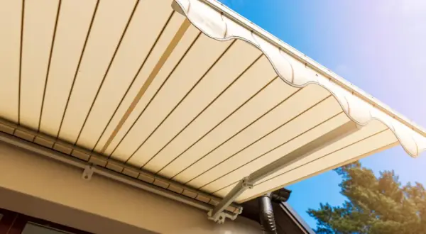 Toldo beige desplegado sobre terraza, cumpliendo la normativa. Cielo azul y árbol al fondo.