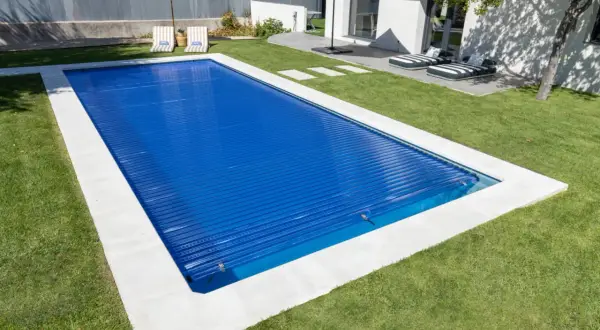 Piscina cubierta con persiana azul, lista para climatizar y disfrutar. Césped verde y tumbonas invitan al relax.