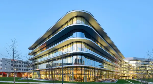 Edificio moderno con **cristales curvos** que reflejan el cielo azul. Fachada ondulada y luminosa, con áreas verdes y árboles alrededor.