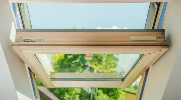 Ventana de tejado abierta, mostrando el marco de madera y el cristal con cámara de aire. Se ve el cielo azul y árboles verdes al exterior.
