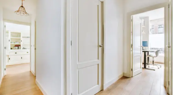Pasillo con puertas de interior blancas y suelo de madera. Se vislumbran una cocina y un despacho.
