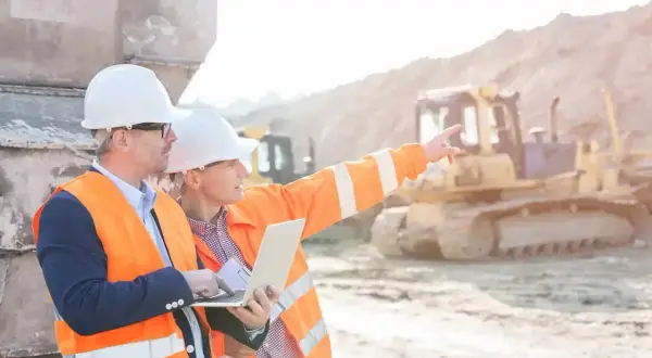 Dos ingenieros con cascos y chalecos naranjas revisan un plano en una obra. Uno señala al fondo, donde se ve una excavadora.