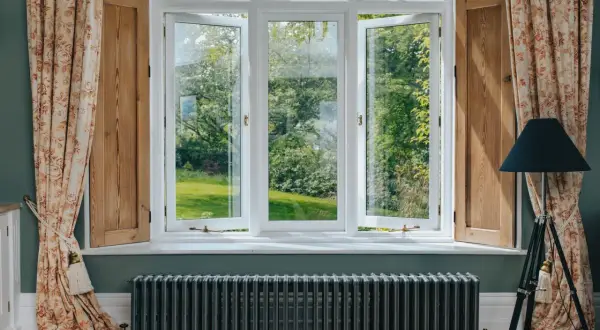 Ventanas blancas abiertas a un jardín verde, con cortinas florales y un radiador antiguo. Las mejores ventanas para disfrutar del exterior.