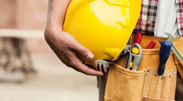 Un trabajador sostiene un casco amarillo. Su cinturón de herramientas tiene destornilladores y una llave inglesa. Esto es lo que es el ITE: seguridad y preparación.