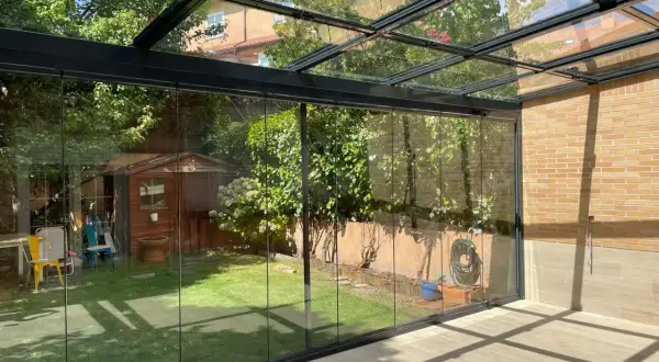 Modernos tejados para porches de cristal, con vistas a un jardín con caseta de madera y árboles frondosos.