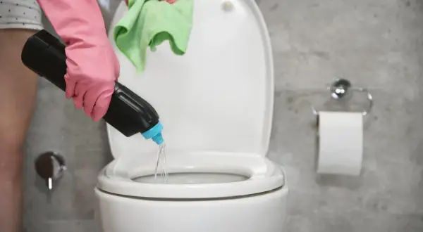 Mano con guante rosa vierte agua fuerte para que sirve en el inodoro. Paño verde en mano.