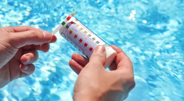 Manos sostienen una tira reactiva para medir el pH de la piscina. El agua azul y cristalina de la piscina se ve al fondo.