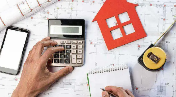 Manos calculando presupuesto para obras de reforma. Planos, calculadora, cinta métrica y casa roja.