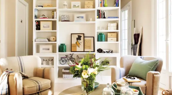 Acogedor salón con estanterías blancas llenas de libros y objetos decorativos, dos sillones de rayas y una mesa de centro con flores. Un rincón que evoca la calidez de las casas pequeñas.