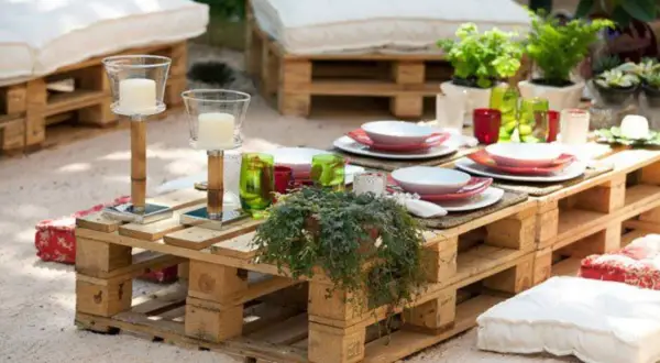 Mesa de jardín con ideas con palets, decorada con vajilla, velas y plantas. Cojines a los lados invitan a relajarse.