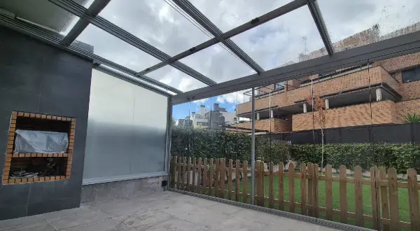 Patio con techo de cristal y estructura metálica, ideal para techar patio económico. Incluye barbacoa y valla de madera.