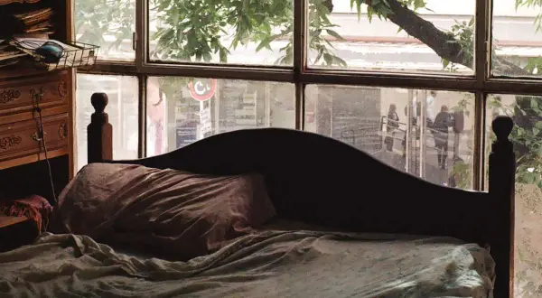 Dormitorio con ventana en el cabecero. La cama está hecha, con sábanas arrugadas y una almohada. A la izquierda, un mueble de madera con cajones.