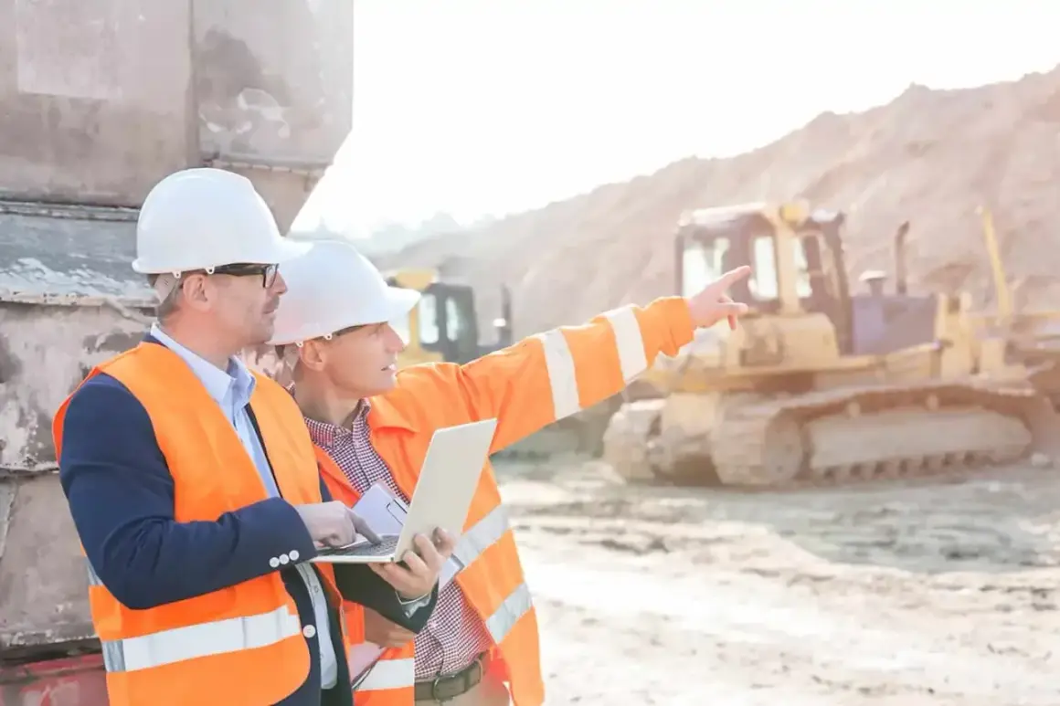Dos ingenieros con cascos y chalecos naranjas revisan un plano en una obra. Uno señala al fondo, donde se ve una excavadora.