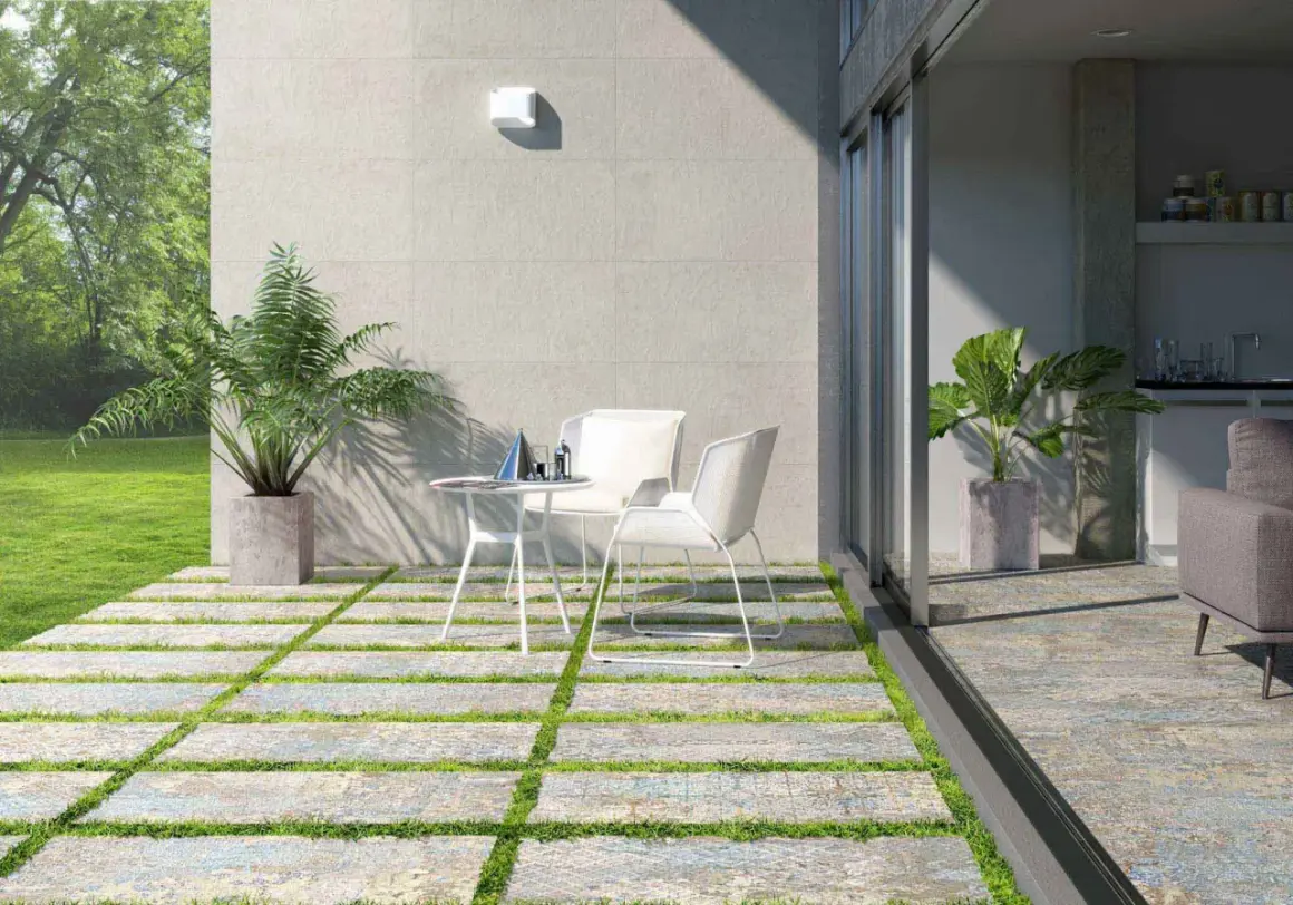 Terrazas pequeñas con encanto, con baldosas y césped, mesa y sillas blancas, plantas y vista a un jardín verde.