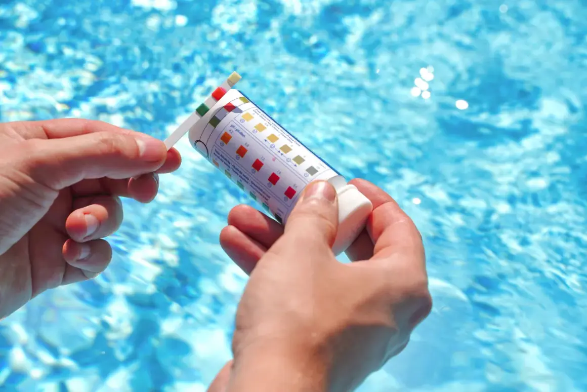 Manos sostienen una tira reactiva para medir el pH de la piscina. El agua azul y cristalina de la piscina se ve al fondo.