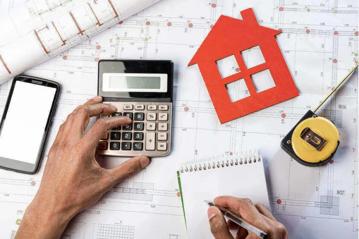 Manos calculando presupuesto para obras de reforma. Planos, calculadora, cinta métrica y casa roja.