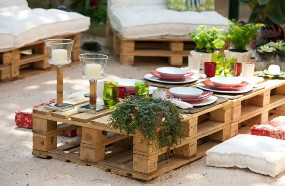 Mesa de jardín con ideas con palets, decorada con vajilla, velas y plantas. Cojines a los lados invitan a relajarse.