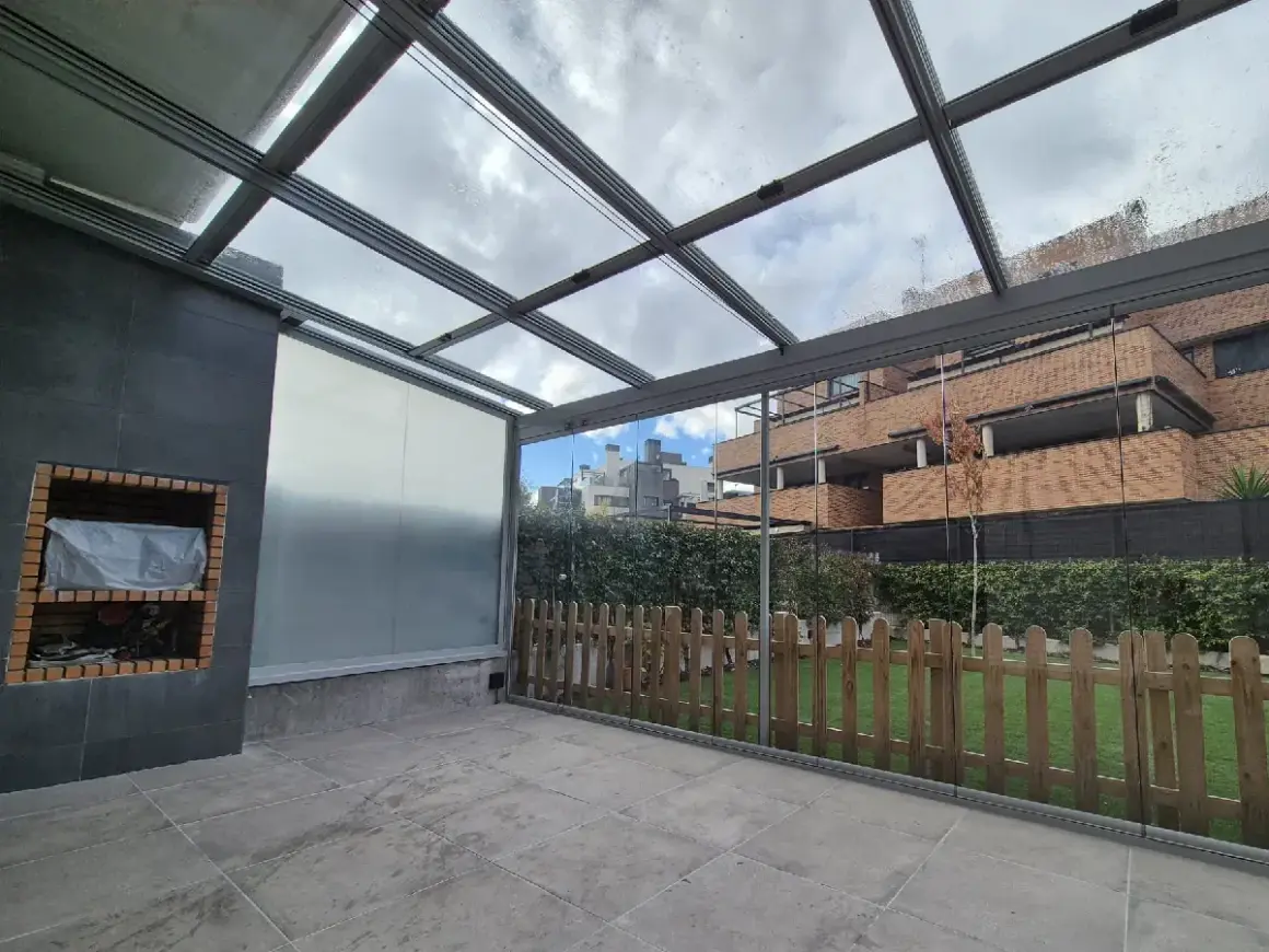 Patio con techo de cristal y estructura metálica, ideal para techar patio económico. Incluye barbacoa y valla de madera.