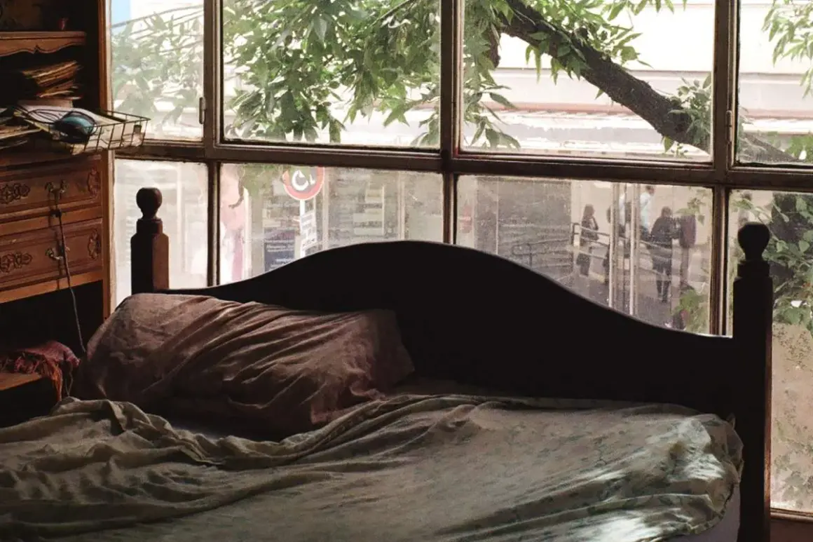 Dormitorio con ventana en el cabecero. La cama está hecha, con sábanas arrugadas y una almohada. A la izquierda, un mueble de madera con cajones.