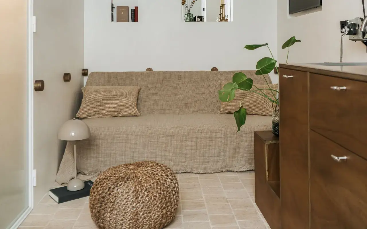 Acogedora habitación de 4 metros cuadrados con sofá cama, puf, planta y mueble de madera.