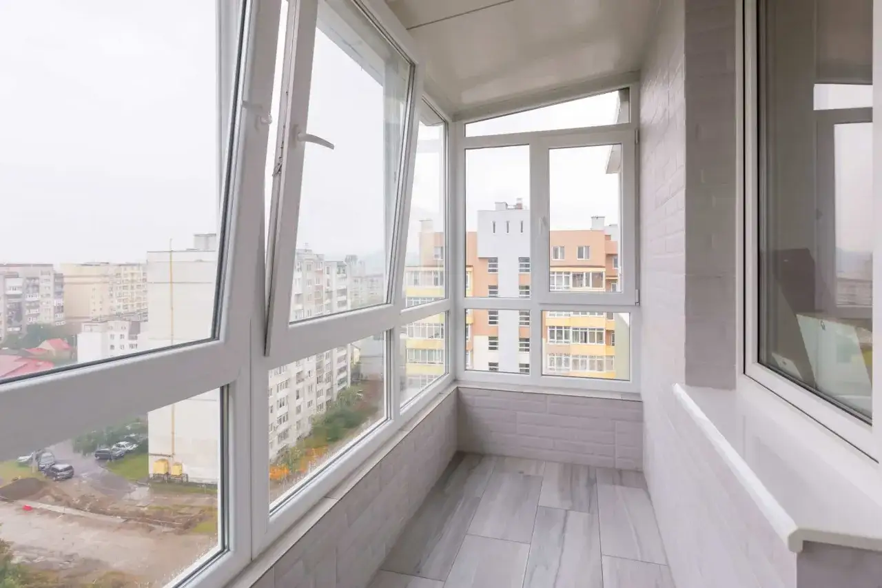 Interior de un balcon cerrado con grandes ventanales que ofrecen vistas a edificios urbanos. El suelo es de baldosas grises y las paredes est&aacute;n revestidas con paneles que imitan ladrillos blancos.