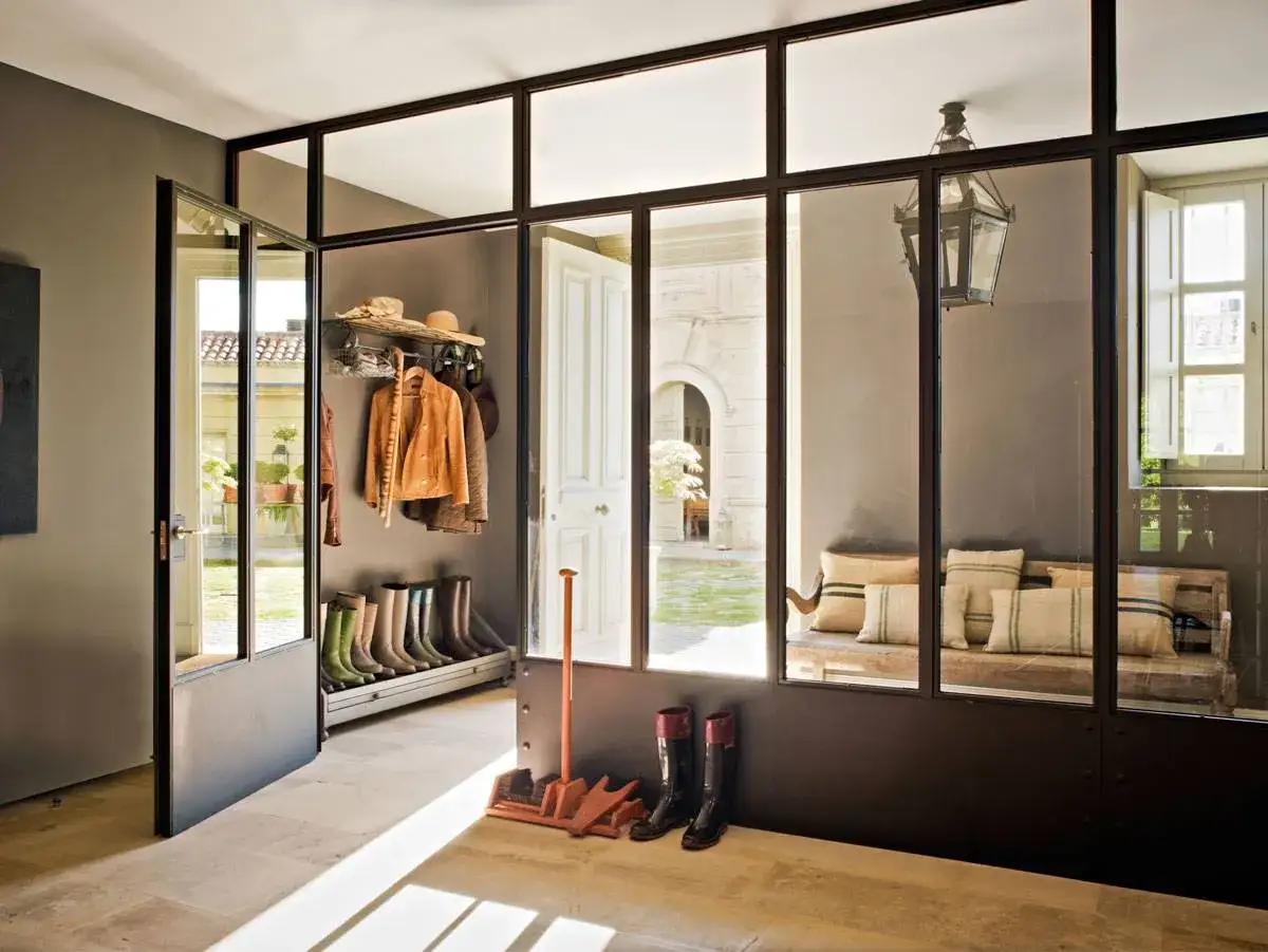 Entrada con botas, chaquetas y sombreros colgados, separada por paredes de cristal de un patio interior con banco y cojines.