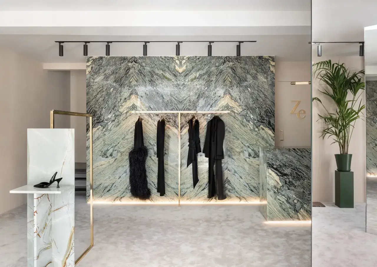 Exhibición de moda con pared de mármol verde veteado, percheros dorados con ropa negra y un zapato de tacón sobre un pedestal de mármol.